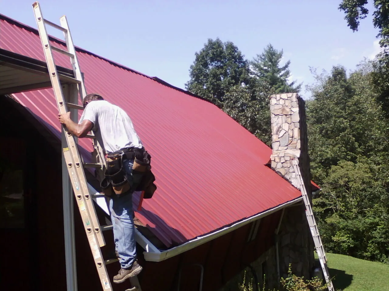 Roofer on a ladder beside a red metal roof and stone chimney — Soffit & Fascia Repair by Roofing Brothers in Schuylerville